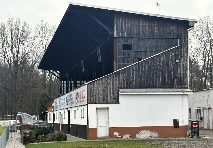 Exteriér historického stadionu v Poděbradech, který je ve špatném stavu, s dřevěnou tribunou a přilehlými budovami.
